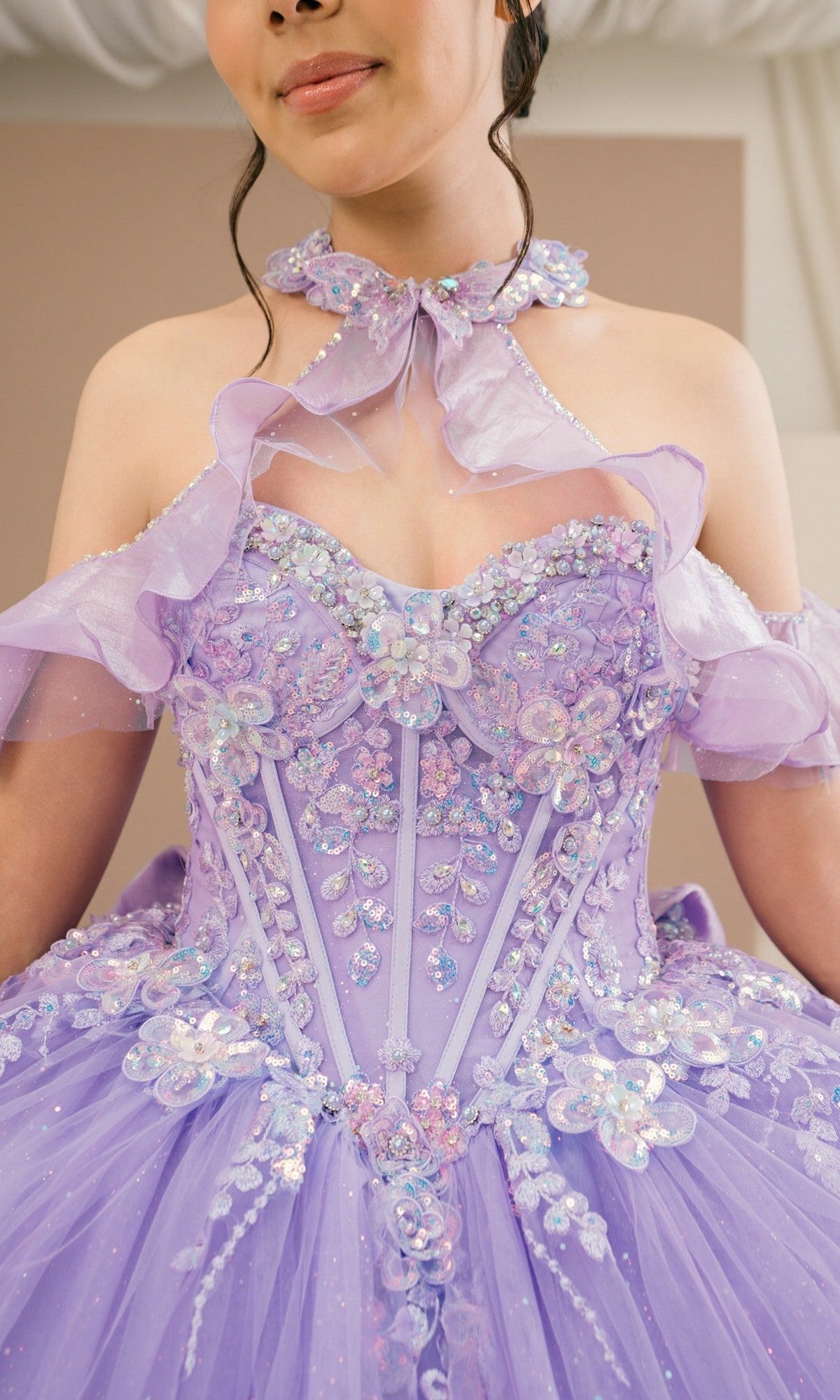 A person wearing the Quinceanera Dress 8585JQ By Cinderella Couture, an ornate lavender ball gown adorned with sequins, floral appliqués, and sheer ruffles, shown from the shoulders to the waist.